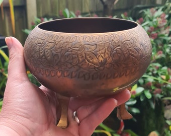 May include: A decorative brass bowl with a floral design and three legs. The bowl is made of a shiny metal and has a warm, golden color.