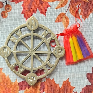 May include: A brass compass with the letters N, E, S, and W around the edge. The compass is on a fabric background with a red, yellow, and purple bag containing candles.