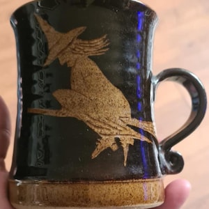 May include: A black ceramic mug with a brown witch silhouette design. The mug has a curved handle and a brown base. The witch is depicted riding a broomstick. The mug is likely handmade and suitable for hot beverages.