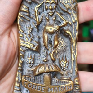 Puede incluir: Un llamador de puerta de latón con forma de duende de pie sobre una seta. El llamador está grabado con "Exmoor Pixie" y "Combe Martin Devon".