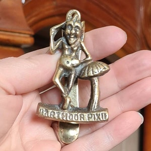 May include: Brass door knocker featuring a whimsical pixie figure holding a mushroom. The knocker is inscribed with the words "Marthour Pixie".