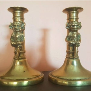 May include: A pair of antique brass candlesticks with a whimsical design. Each candlestick features a small, detailed figure of a mythical creature with horns and a beard, sitting on a column.