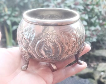 May include: A silver, ornate, footed bowl with an intricate floral design. The bowl has a round shape and three legs.
