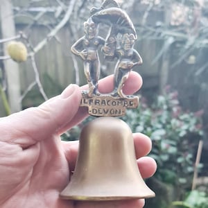 Może przedstawiać: Dzwonek mosiężny z ozdobnym wierzchołkiem przedstawiającym dwie postacie trzymające parasol. Dzwonek jest wygrawerowany napisem "Ilfracombe Devon".