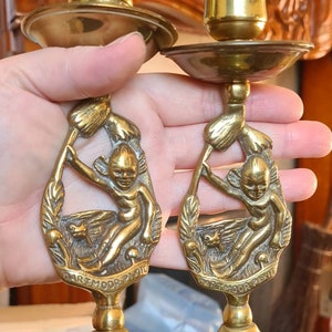 May include: Two brass candle holders with a whimsical design. Each holder features a small fairy-like figure holding a mushroom. The base of each holder is inscribed with "Dartmoor Pixie".