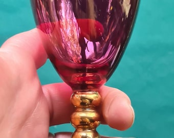 May include: A small, clear glass goblet with a purple hue and gold trim. The goblet has a round bowl and a stem with three gold beads. The goblet is sitting on a table.