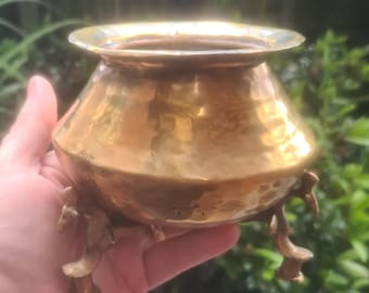 May include: A small, hammered brass pot with a wide rim and three decorative feet. The pot has a rounded body and a warm, golden color. The surface of the pot has a textured, dimpled appearance.