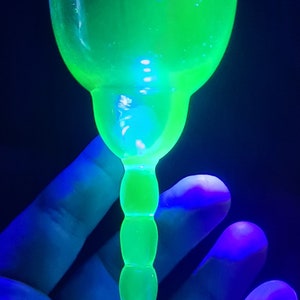 Può includere: Un calice di vetro verde con un gambo scanalato. Il calice è illuminato da una luce nera, che lo fa brillare.