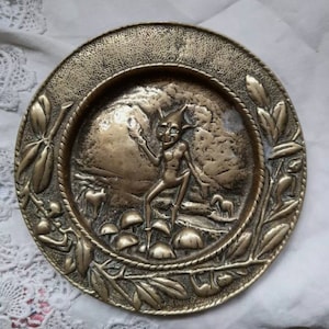 Könnte beinhalten: Eine Messingplatte mit einem erhabenen Design eines Kobolds oder Kobolds, der auf einem Pilz steht. Der Kobold hält ein kleines Objekt in seiner Hand. Die Platte ist von einem Rand aus Blättern und Pilzen umgeben.