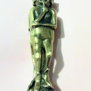 May include: A bronze-colored figurine of a smiling, humanoid figure with pointed ears, holding a finger to its lips. The figure is standing on a base with engraved text. The figurine has a loop at the top for hanging.