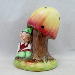 May include: A ceramic figurine of a sleeping gnome leaning against a yellow and red mushroom with brown spots. The gnome is wearing a green hat, red waistcoat, and green trousers.