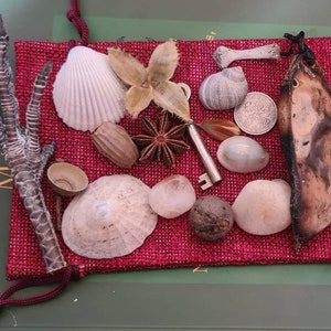 Może przedstawiać: Czerwona materiałowa torebka wypełniona różnymi małymi przedmiotami, w tym ptasim pazurem, muszelkami, kluczem, anyżem gwiazdkowym, monetą i suchą łuszczyną grochu.