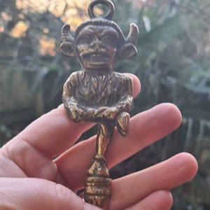 Puede incluir: Una llave de color latón con una figura detallada de un diablo sonriente con cuernos y un lazo. La llave tiene un diseño ornamentado y se sostiene contra un fondo borroso al aire libre.
