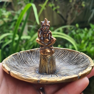 Puede incluir: Un porta anillos de latón con forma de hongo con una pequeña figura detallada de una criatura sentada en la parte superior. La figura tiene un sombrero puntiagudo y una cara detallada.