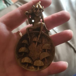Op de afbeelding: Een bronzen metalen beeldje van een goblin die op een ronde schijf zit met een reliëf van paddenstoelen. De schijf heeft de woorden "GOOD LUCK" en "PARKER" erop gegraveerd.