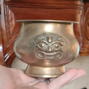 May include: A brass bowl with a decorative design of a face with a third eye. The bowl has a wide rim and a rounded base.