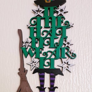 May include: Green wooden witch cutout with the text "If the Hat Fits Wear It" and a brown broom. The witch is wearing a black hat, purple and white striped stockings, and black shoes with gold buckles.