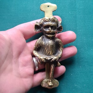 May include: A brass door knocker shaped like a monkey with a mischievous expression. The monkey is sitting with its legs crossed and holding a small object in its hands. The knocker is attached to a rectangular base with a decorative top.