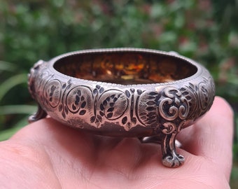 May include: A silver salt cellar with an ornate design and three legs. The cellar has a circular shape and is decorated with a floral pattern.