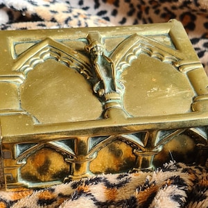 Puede incluir: Una caja de latón con un diseño ornamentado con arcos y una figura. La caja tiene una superficie texturizada y un color dorado.