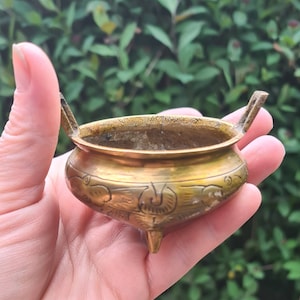 May include: A small, gold-colored brass incense burner with intricate floral designs. The burner has three legs and two handles.