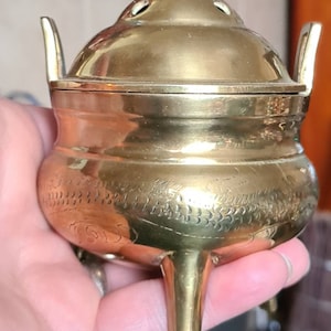 May include: A small, gold-colored brass incense burner with a lid and three legs. The burner has a decorative design of stylized flowers and leaves.