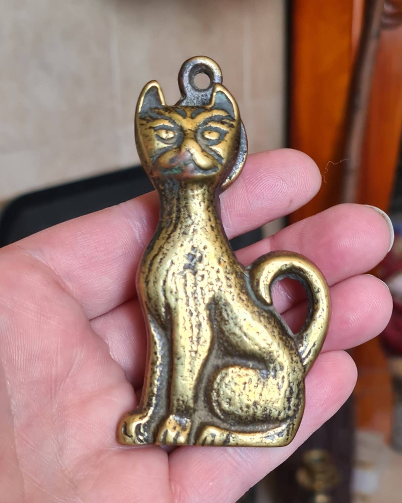 May include: A brass cat figurine with a detailed face and a textured surface. The cat is sitting with its tail curled around its body.