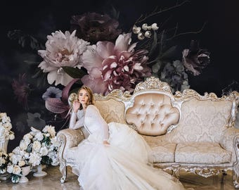 May include: A long-sleeved white gown with a full skirt sits on an ornate gold-framed sofa with beige tufted upholstery. The background features a large-scale mural of dark, moody floral arrangements including pink and white peonies and lilies.