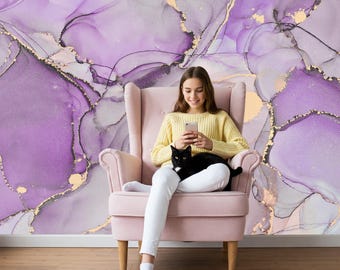May include: A light pink upholstered armchair with high wings and wooden legs. The chair features a smooth fabric finish and a structured backrest. It is positioned against a wall covered in wallpaper with a purple and gold marble pattern.