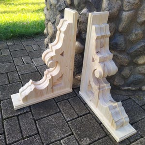 Può includere: Due mensole in legno non rifinite con disegni intagliati ornati. Le mensole sono di colore chiaro e presentano un motivo decorativo a volute, perfette per aggiungere un tocco di eleganza a qualsiasi progetto di arredamento per la casa.