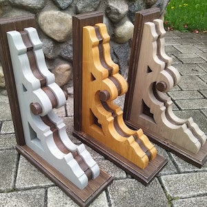 May include: Three decorative wooden corbels in varying colours: grey and brown, golden oak and brown, and natural wood and brown. Each corbel features an ornate, scroll-like design and is mounted on a rectangular base. The corbels are displayed on a stone surface.