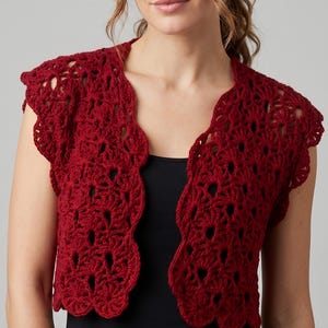 May include: A deep red crochet bolero featuring an intricate openwork fan pattern with a scalloped edge. This short-sleeved garment has an open front design and is worn over a plain black top, showcasing the detailed texture of the yarn work.