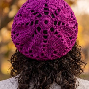 Può includere: Un basco all'uncinetto color magenta vivace caratterizzato da un motivo traforato circolare a forma di stella che si irradia dal centro. Il cappello è realizzato con filato spesso, mostrando un design testurizzato con spazi vuoti.