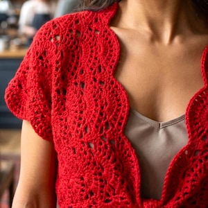 May include: A vibrant red, open-front crochet shrug with a scalloped edge. The short-sleeved bolero style features an intricate openwork design and a tie closure at the front. The garment is made of a soft, textured yarn.