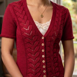 May include: A deep red short-sleeved knit cardigan featuring a V-neckline and a vertical leaf-patterned lace design along the front panels. The garment is secured with a row of seven light-colored buttons and is worn over a cream-colored camisole.