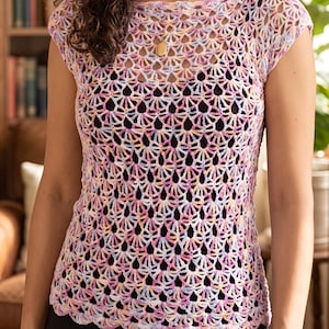 Puede incluir: Una blusa de ganchillo de manga corta con un delicado patrón de abanicos calados en tonos pastel variados de rosa, amarillo y azul claro. La prenda tiene un escote barco y un dobladillo festoneado, usada sobre una capa base negra.
