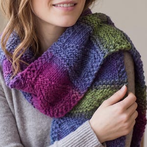 Könnte beinhalten: Ein grob gestrickter Schal mit einem bunten Farbmuster aus tiefem Violett, Magenta und Olivgrün. Der Schal ist aus dickem, strukturiertem Garn im klassischen Kraus-Rechts-Muster gefertigt und erzeugt ein weiches, voluminöses Aussehen mit Farbblöcken.