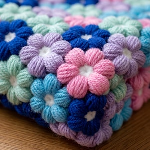 Puede incluir: Una manta de ganchillo hecha de motivos florales interconectados en tonos rosa, lavanda, azul claro y azul oscuro. Cada flor presenta ocho pétalos redondeados que rodean un centro blanco, creando un patrón texturizado y repetitivo.