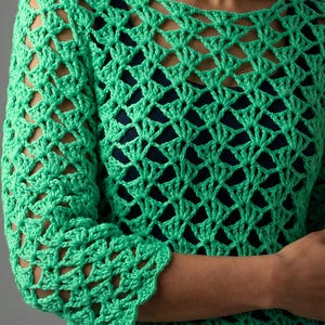 May include: A bright green long-sleeve crochet top featuring an openwork shell stitch pattern. The garment has a wide neckline and is layered over a solid dark navy blue base layer, showcasing the intricate texture of the yarn construction.