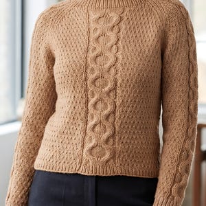 Könnte beinhalten: Ein beigefarbener Langarmpullover mit einem zentralen vertikalen Zopfmuster, das von strukturierten Wabenstrickbahnen flankiert wird. Das Kleidungsstück hat einen gerippten Rundhalsausschnitt, gerippte Bündchen und einen gerippten Saum aus hellbraunem Garn.