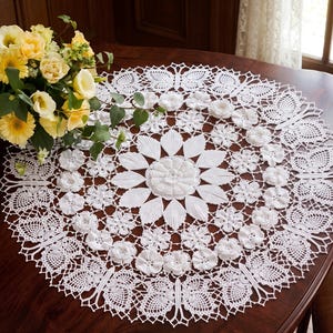 May include: A white, round, crocheted doily with a floral design. The center features a large flower, surrounded by smaller flowers and intricate lace patterns. The doily is placed on a dark wooden table, with a bouquet of yellow flowers in the background.