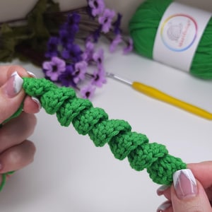 May include: A close-up of a green crocheted chain with a series of small, round bumps. The chain is being held by a person's hands.