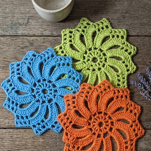 May include: Three circular crochet coasters in bright blue, lime green, and orange, each featuring a radiating spiral pattern with openwork details. The coasters are arranged in a cluster alongside a speckled ceramic mug and lavender sprigs.
