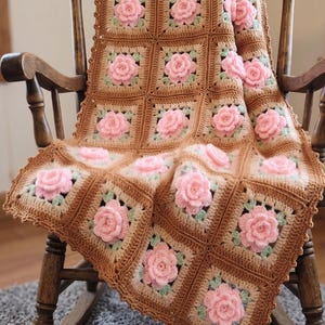 May include: A handmade crochet blanket composed of square panels featuring three-dimensional light pink roses with small green leaves, set against a tan and brown yarn background with a scalloped border edge throughout the entire textile piece.