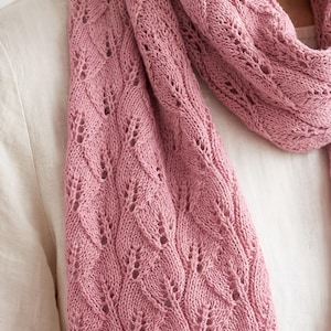 May include: A dusky pink knitted scarf featuring a detailed openwork leaf design. The scarf is draped, highlighting the intricate pattern and soft texture of the yarn. A light and airy accessory.