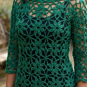 May include: A dark green crochet top featuring an intricate, repeating floral motif pattern throughout the garment. The piece has three-quarter length sleeves, a wide neckline, and is worn over a solid dark-colored base layer garment.