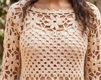 Op de afbeelding: Een beige top met lange mouwen gemaakt van ingewikkeld haakwerk met een herhalend bloemmotief over het hele kledingstuk. Het ontwerp heeft een wijde halslijn en een semi-transparante constructie die een lichtgekleurde basislaag eronder onthult.