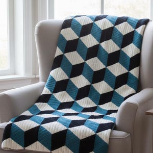 Pode incluir: Uma manta de malha com um padrão geométrico de cubo em azul-petróleo, preto e branco, drapeada sobre uma poltrona cinza. O design da manta cria um efeito 3D. A poltrona está perto de uma janela.