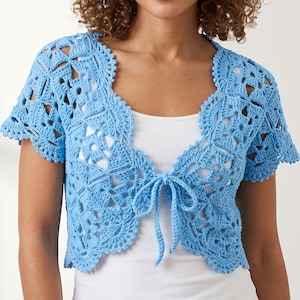 Può includere: Un bolero all'uncinetto azzurro caratterizzato da un intricato motivo traforato ispirato ai fiori, maniche corte e una chiusura con laccetti frontali. Il capo è indossato sopra una canotta bianca semplice, mettendo in risalto i dettagli e i bordi smerlati.