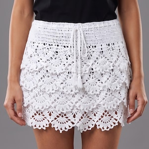 Puede incluir: Una minifalda de ganchillo blanca con un diseño de capas, bordes festoneados y una cintura con cordón ajustable y dos borlas. La falda está confeccionada con hilo blanco en un patrón floral detallado, creando un aspecto superpuesto con dobladillo.